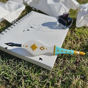 May include: A white and light blue dip pen with gold star and diamond accents, featuring a metal nib. The pen rests on a spiral-bound sketchbook containing a hand-drawn circular sketch and ink splatters, accompanied by a small glass ink bottle.