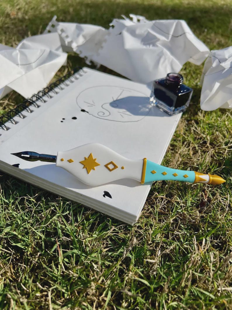 May include: A white and light blue dip pen with gold star and diamond accents, featuring a metal nib. The pen rests on a spiral-bound sketchbook containing a hand-drawn circular sketch and ink splatters, accompanied by a small glass ink bottle.