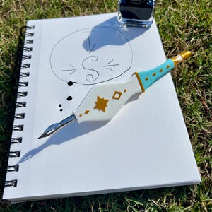 May include: A white and light blue dip pen with gold star and diamond accents, featuring a metal nib. The pen rests on a spiral-bound notebook containing a hand-drawn circle with the letter S inside, accompanied by a small glass ink bottle.