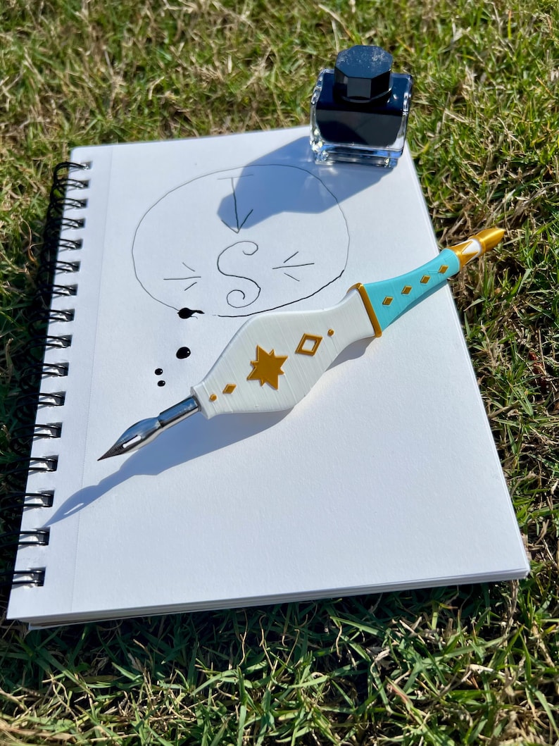 May include: A white and light blue dip pen with gold star and diamond accents, featuring a metal nib. The pen rests on a spiral-bound notebook containing a hand-drawn circle with the letter S inside, accompanied by a small glass ink bottle.