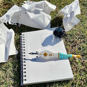 May include: A white spiral-bound sketchbook with a hand-drawn circle and arrow, a dip pen with a white, gold, and turquoise handle, and a small square glass inkwell. Several crumpled pieces of white paper are scattered around the sketchbook.