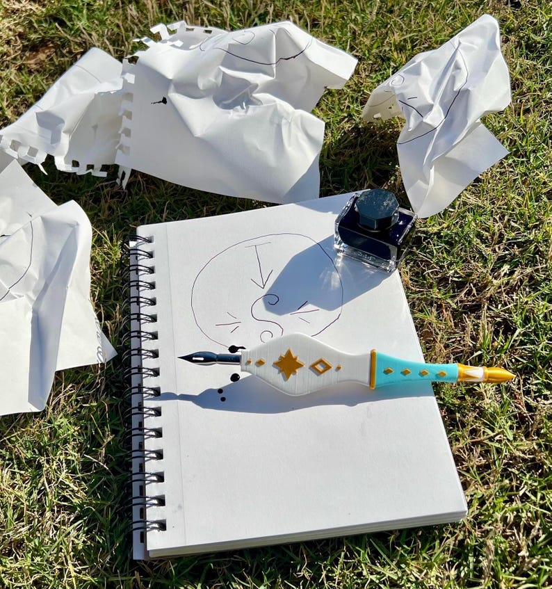 May include: A white spiral-bound sketchbook with a hand-drawn circle and arrow, a dip pen with a white, gold, and turquoise handle, and a small square glass inkwell. Several crumpled pieces of white paper are scattered around the sketchbook.