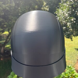 Peut inclure: Un casque en plastique noir avec un dessus arrondi lisse et une visi&egrave;re plate. Le casque a une finition mate et semble &ecirc;tre imprim&eacute; en 3D.