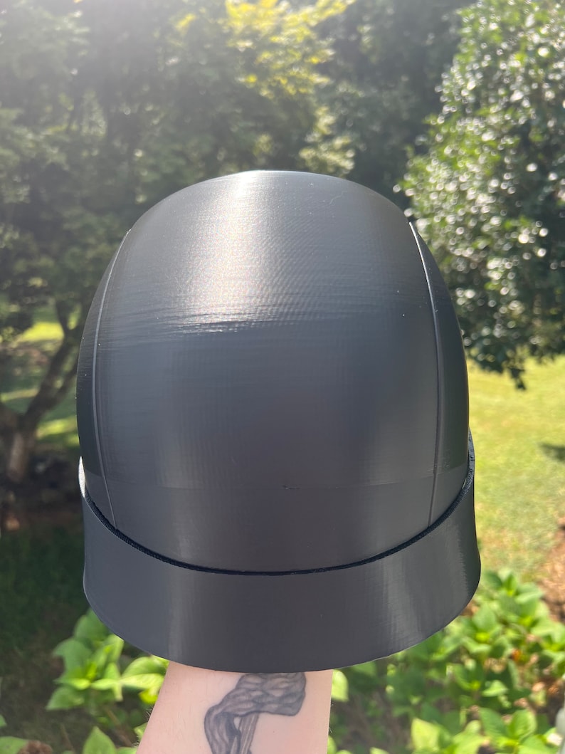 Peut inclure: Un casque en plastique noir avec un dessus arrondi lisse et une visi&egrave;re plate. Le casque a une finition mate et semble &ecirc;tre imprim&eacute; en 3D.