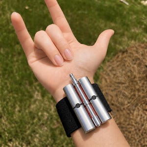 May include: A black elastic wristband featuring two cylindrical silver metal components attached to the front. Each silver cylinder has a small black hexagonal detail at the centre, and a thin silver rod is positioned between the two cylinders.
