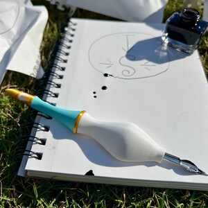 May include: A dip pen with a white bulbous grip, a turquoise midsection, and a gold ring accent, resting on a spiral-bound sketchbook. The sketchbook contains a hand-drawn circular logo with the letter S and a T, alongside a small glass inkwell.