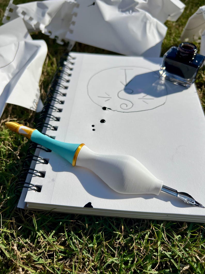 May include: A dip pen with a white bulbous grip, a turquoise midsection, and a gold ring accent, resting on a spiral-bound sketchbook. The sketchbook contains a hand-drawn circular logo with the letter S and a T, alongside a small glass inkwell.