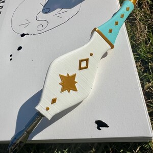 May include: A dip pen with a white, bulbous handle featuring gold star and diamond-shaped accents. The handle transitions to a light blue grip with gold diamond details and a gold-colored tip. A metal nib is attached to the bottom of the handle.