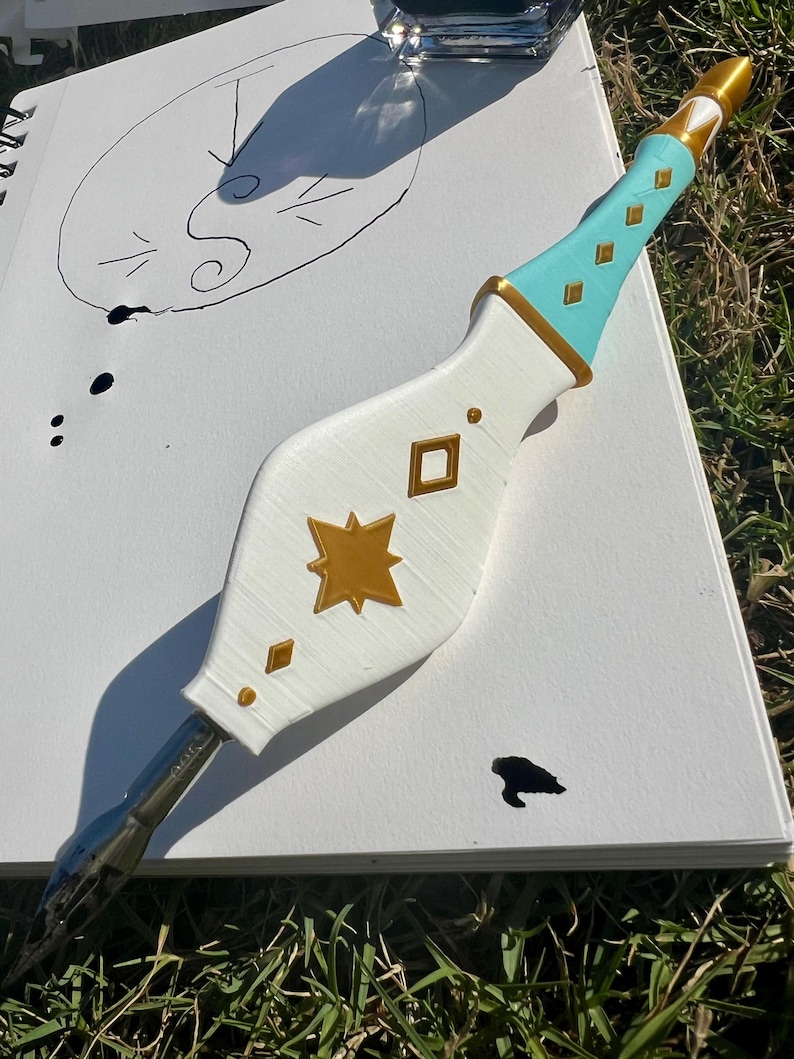 May include: A dip pen with a white, bulbous handle featuring gold star and diamond-shaped accents. The handle transitions to a light blue grip with gold diamond details and a gold-colored tip. A metal nib is attached to the bottom of the handle.