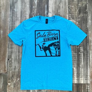 Pu&ograve; includere: Una t-shirt blu turchese con una stampa grafica nera di una mucca in un recinto con il testo "Sale Barn Reject".