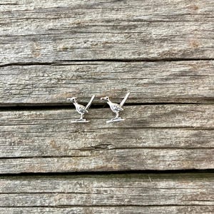 May include: A pair of silver-colored roadrunner bird earrings. The earrings are small, detailed, and feature the bird's distinctive upright posture and long tail. The earrings are set against a rustic, weathered wooden background.