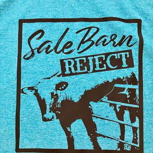 Pu&ograve; includere: Una t-shirt blu turchese con una grafica nera di una mucca in un recinto. La grafica contiene il testo "Sale Barn Reject" in uno striscione nero.