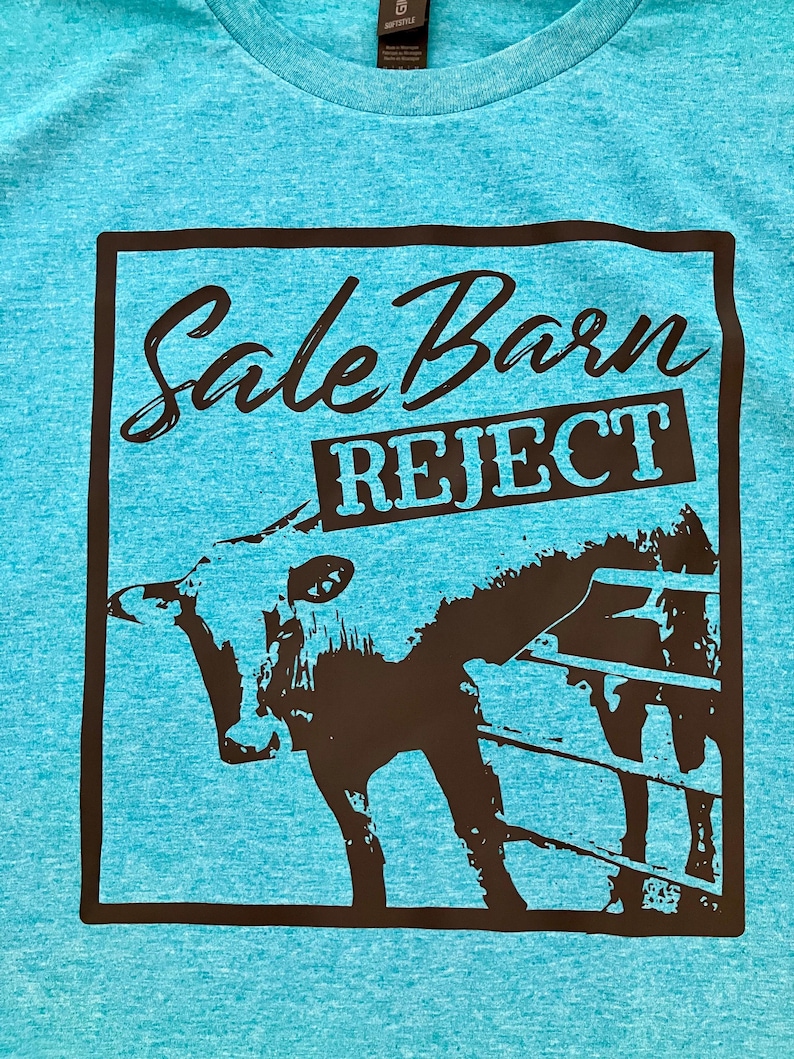 Pu&ograve; includere: Una t-shirt blu turchese con una grafica nera di una mucca in un recinto. La grafica contiene il testo "Sale Barn Reject" in uno striscione nero.