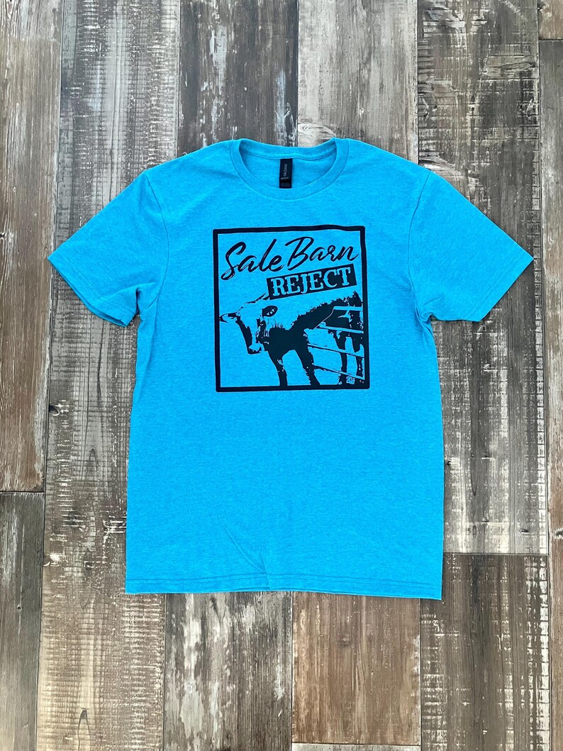 Pu&ograve; includere: Una t-shirt blu turchese con una stampa grafica nera di una mucca in un recinto con il testo "Sale Barn Reject".