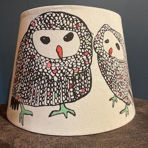 May include: A tapered drum lampshade with an off-white background featuring a black line-art illustration of two owls. The owls have orange beaks, green feet, and scattered pink and green dots within their black-outlined, textured feather patterns.