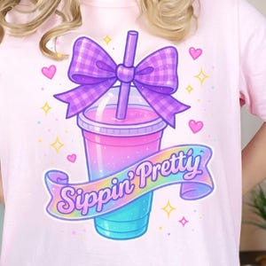 Könnte beinhalten: Pinkes T-Shirt mit einem Grafikdruck eines bunten Getränks mit einer lila Schleife und dem Text "Sippin' Pretty".