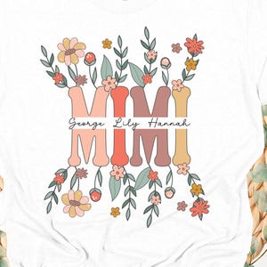 Peut inclure: T-shirt blanc avec le mot "MIMI" dans un motif floral. Les noms "George Lily Hannah" sont imprimés sous le mot "MIMI". Le t-shirt a un col rond et des manches courtes. Le motif comprend des fleurs dans les tons de rose, d'orange et de jaune.
