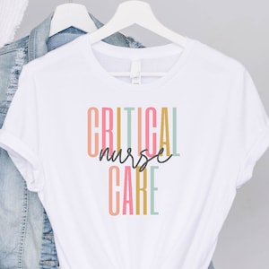 May include: White t-shirt with the text "CRITICAL nurse CARE" in colorful vertical stripes. The text is in a rainbow color scheme with a pink, green, yellow, and blue color palette.