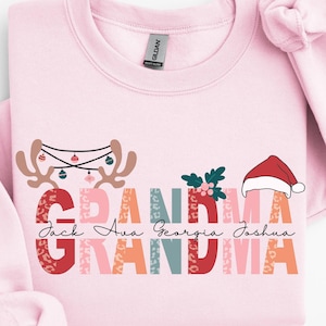 May include: A light pink sweatshirt with the word "Grandma" in a colorful, patterned design. The design includes reindeer antlers, holly, and a Santa hat. Names are printed below the word Grandma.