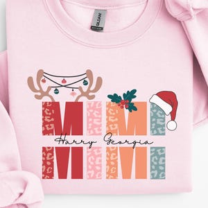 Peut inclure: Un sweat-shirt rose clair avec un motif festif. Le motif comprend le mot "MIMI" dans un style de lettres en blocs, des bois de rennes, un bonnet de Père Noël et les noms "Harry Georgia". Le sweat-shirt est en matière douce.