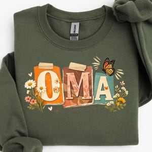 May include: An olive green sweatshirt with the word "OMA" in a colorful, floral design. The letters are on individual rectangles with a butterfly and flowers. The sweatshirt is made of a soft material.