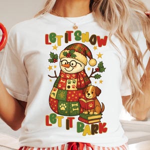 May include: White t-shirt with a festive design. It features a snowman and a dog, with the text "Let It Snow, Let It Bark". The snowman wears a plaid hat and scarf, and the dog reads a book. Holly and stars are also included.
