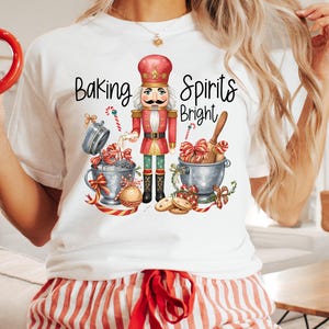 Könnte beinhalten: Weißes T-Shirt mit einem farbenfrohen Grafikdesign eines Nussknackers, Zuckerstangen und dem Text "Baking Spirits Bright". Die Person trägt eine rot-weiß gestreifte Pyjamahose und hält einen Lebkuchenmann-Becher.