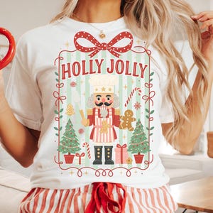 May include: White t-shirt with a "HOLLY JOLLY" graphic featuring a nutcracker, Christmas trees, and gingerbread men. The person is also wearing red and white striped pajama pants. A gingerbread man mug is held in the left hand.