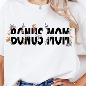 May include: White t-shirt with black text that reads "Bonus Mom" with the names "Archer, Charlie, Jasmine" written in smaller font underneath. The text is surrounded by colourful floral designs.