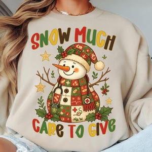 Könnte beinhalten: Beiges Sweatshirt mit einem farbenfrohen Design eines Schneemanns, der einen medizinisch thematisierten Schal und Hut trägt. Der Text "SNOW MUCH CARE TO GIVE" ist oberhalb und unterhalb der Schneemann-Illustration angeordnet. Der Schneemann hat eine Karottennase.