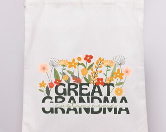 Op de afbeelding: Een witte draagtas met een bloemenontwerp en de woorden "GREAT GRANDMA" in een vetgedrukte lettertype. Het bloemenontwerp bevat oranje, gele en rode bloemen. De woorden "custom name" staan eronder.