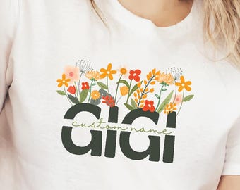 May include: White t-shirt with a floral design and the name "Gigi" in dark green. The words "custom name" are above the name. The shirt has a crew neck and short sleeves. The model is wearing blue jeans and a brown jacket.