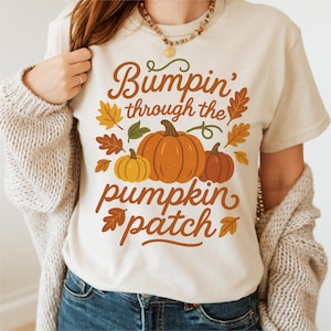 May include: Cream-colored t-shirt with the text "Bumpin' through the pumpkin patch" in brown script. The design includes orange, yellow, and brown pumpkins and fall leaves. A cozy fall-themed graphic tee.
