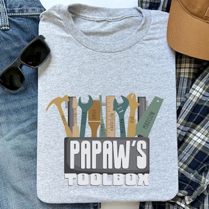 May include: A light gray t-shirt featuring a toolbox design with various tools and names. The text "PAPAW'S TOOLBOX" is prominently displayed. The shirt is surrounded by sunglasses, a baseball cap, and denim.