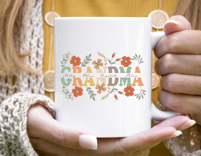 May include: White ceramic mug with the word "GRANDMA" in retro orange, green, and tan lettering, adorned with floral accents and the names Molly, Ethan, Elizabeth, and Miles. The mug has a white handle.