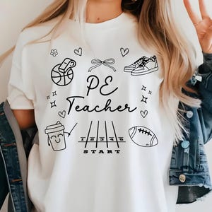 Puede incluir: Camiseta blanca con ilustraciones en negro de equipamiento deportivo y la frase "PE Teacher". El diseño incluye una pelota de baloncesto, zapatillas deportivas, un balón de rugby y una taza de café.