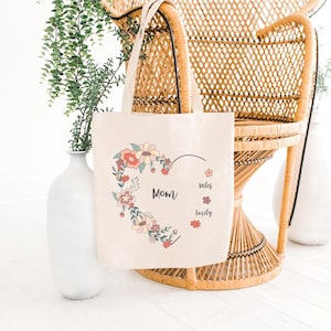 Pode incluir: Uma bolsa de lona bege com um design de cora&ccedil;&atilde;o floral. O cora&ccedil;&atilde;o &eacute; contornado em preto e preenchido com flores rosa, amarelas e verdes. O texto "Mom" est&aacute; dentro do cora&ccedil;&atilde;o, e os nomes "Miles" e "Emily" est&atilde;o escritos abaixo do cora&ccedil;&atilde;o.