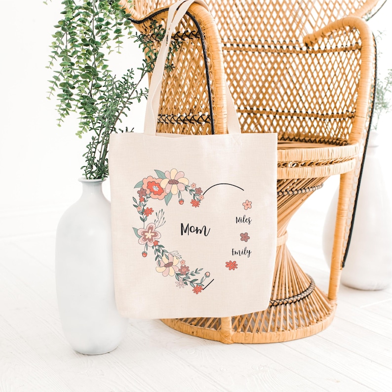 Pode incluir: Uma bolsa de lona bege com um design de cora&ccedil;&atilde;o floral. O cora&ccedil;&atilde;o &eacute; contornado em preto e preenchido com flores rosa, amarelas e verdes. O texto "Mom" est&aacute; dentro do cora&ccedil;&atilde;o, e os nomes "Miles" e "Emily" est&atilde;o escritos abaixo do cora&ccedil;&atilde;o.