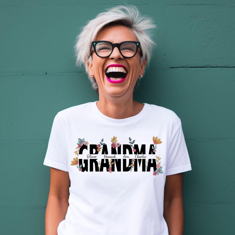 May include: White t-shirt with the word "Grandma" in black letters with floral accents. The names "Oliver, Hannah, Ava, Charlie" are written in smaller letters above the word "Grandma".