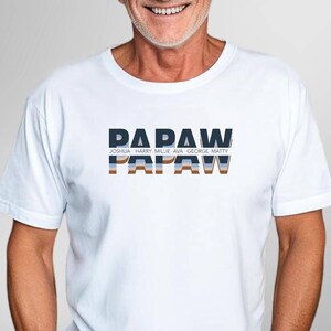 May include: White t-shirt with the word "PAPAW" in a blue and brown design. Below the word "PAPAW" are the names Joshua, Harry, Millie, Ava, George, and Matty. A second "PAPAW" is below the first.