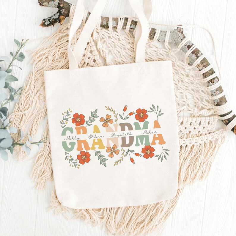 May include: Cream-colored tote bag with the word "GRANDMA" in colorful, retro-style letters, surrounded by floral illustrations. Below the word "GRANDMA" are the names Molly, Ethan, Elizabeth, and Miles. The bag has cream-colored handles.