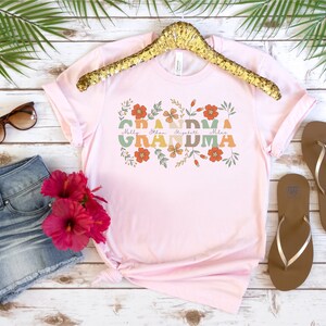 May include: A light pink t-shirt with the word "Grandma" in a floral design. The names Molly, Ethan, Elizabeth, and Miles are printed below the word "Grandma". The shirt is displayed with denim shorts, sunglasses, and sandals.