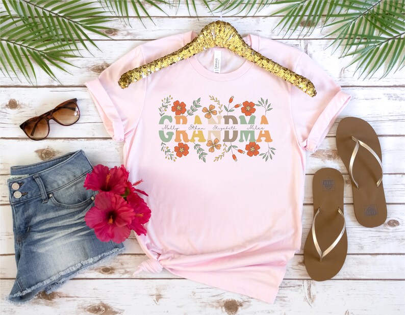 May include: A light pink t-shirt with the word "Grandma" in a floral design. The names Molly, Ethan, Elizabeth, and Miles are printed below the word "Grandma". The shirt is displayed with denim shorts, sunglasses, and sandals.