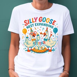 May include: White t-shirt with a colorful graphic featuring two cartoon geese holding baby bottles, wearing party hats and glasses. The text reads "SILLY GOOSE NEST EXPANDING" with "NEW ARRIVAL!" on a banner below the geese.