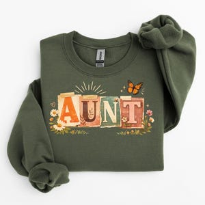 Puede incluir: Una sudadera verde oliva con la palabra "AUNT" en letras coloridas de estilo vintage. El diseño incluye una mariposa, flores y rayos de sol, creando una estética encantadora y caprichosa. Una prenda cómoda e informal.