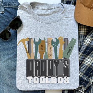 Könnte beinhalten: Hellgraues T-Shirt mit dem Text "DADDY'S TOOLBOX" und Werkzeugillustrationen. Die Werkzeuge umfassen einen Hammer, einen Schraubenschlüssel, einen Pinsel, ein Lineal und eine Säge, jeweils mit einem Namen. Sonnenbrille und Baseballkappe sind ebenfalls sichtbar.