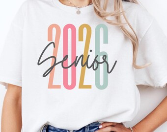 Peut inclure: T-shirt blanc avec le texte "Senior 2026" en lettres majuscules colorées, verticales et en blocs. Le mot "Senior" est écrit en cursive noire.