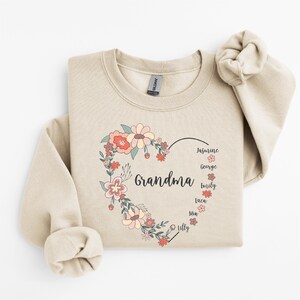 Pode incluir: Uma camisola bege com um design de cora&ccedil;&atilde;o floral. O cora&ccedil;&atilde;o diz "Av&oacute;" e &eacute; rodeado por nomes: Jasmine, George, Emily, Luca, Mia e Lilly.