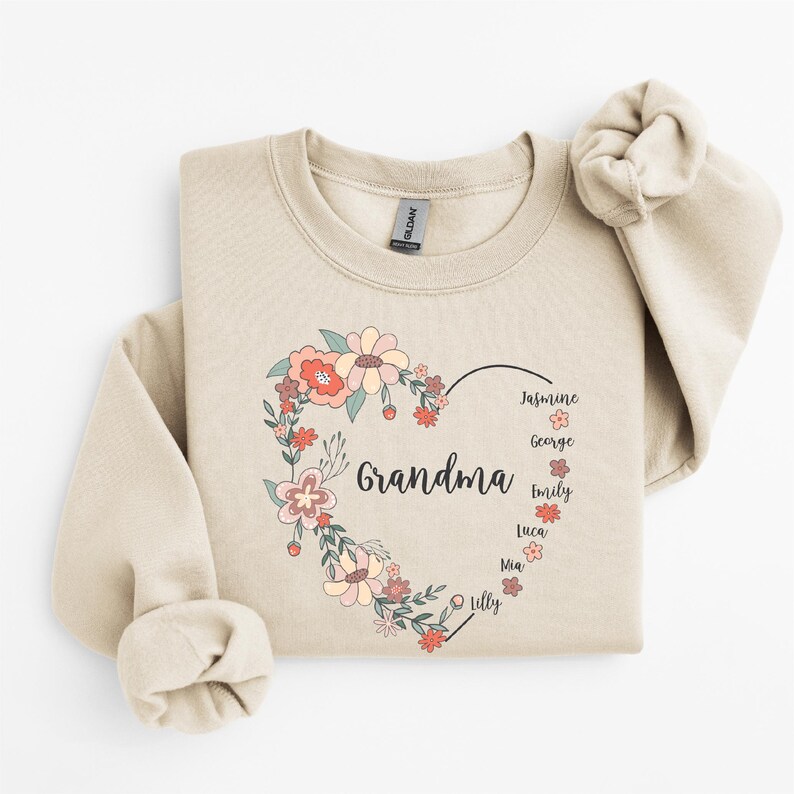 Pode incluir: Uma camisola bege com um design de cora&ccedil;&atilde;o floral. O cora&ccedil;&atilde;o diz "Av&oacute;" e &eacute; rodeado por nomes: Jasmine, George, Emily, Luca, Mia e Lilly.