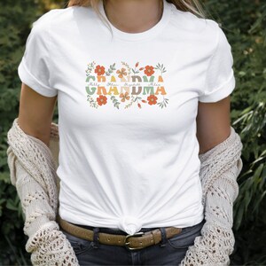May include: White t-shirt with the word "GRANDMA" in colorful, retro-style lettering, surrounded by floral designs. Below the word "GRANDMA" are the names: Milly, Ethan, Elizabeth, and Mila. The shirt is a casual, everyday wear item.